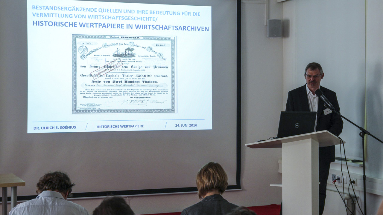 Dr. Ulrich S. Soénius steht am Pult, hinter ihm seine Präsentation über historische Wertpapiere.