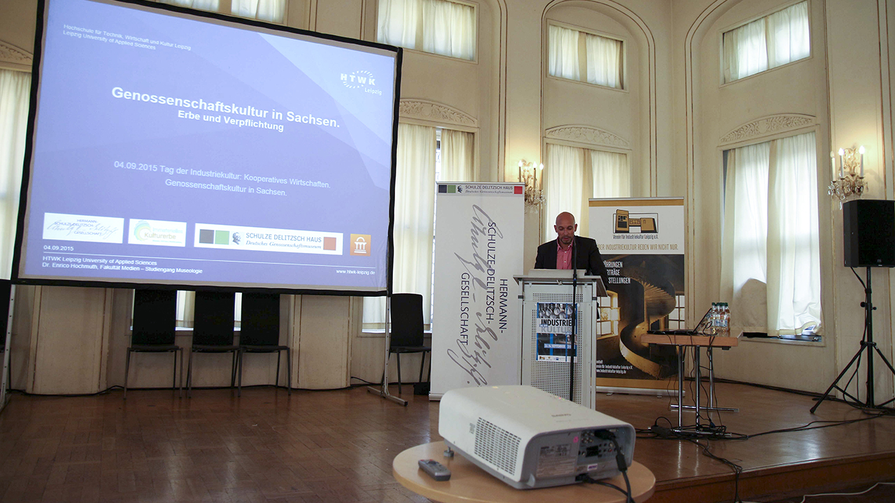 Dr. Enrico Ruge steht am Pult in der Alten Handelsbörse Leipzig, hinter ihm die Präsentation zur Genossenschaftskultur in Sachsen.